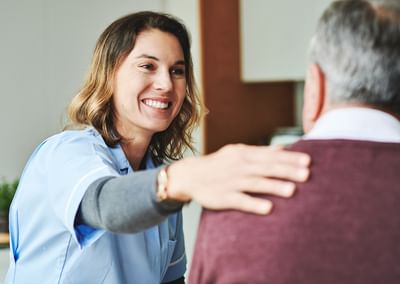 Smiling caregiver in blue scrubs greets an older person, hand on their shoulder.