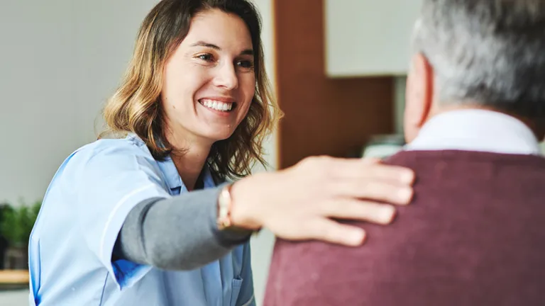 Smiling caregiver in blue scrubs greets an older person, hand on their shoulder.