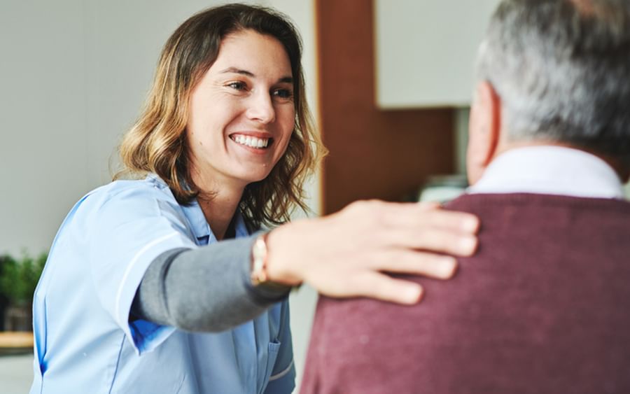 Smiling caregiver in blue scrubs greets an older person, hand on their shoulder.