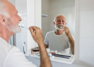 Older person with white hair and beard brushing teeth in a bathroom, smiling in the mirror