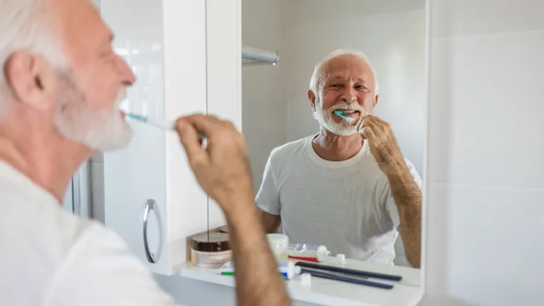 Older person with white hair and beard brushing teeth in a bathroom, smiling in the mirror