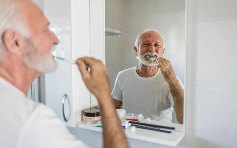Older person with white hair and beard brushing teeth in a bathroom, smiling in the mirror