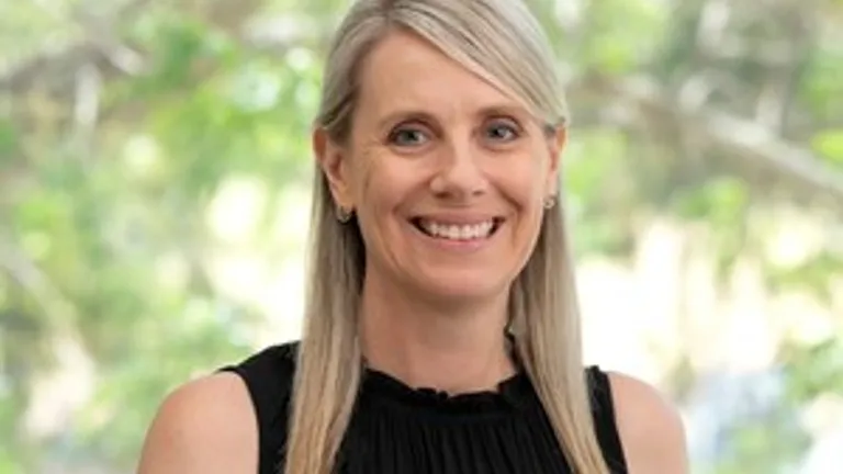 Person with shoulder-length blonde hair, black sleeveless top, yellow flower pin, smiling outdoors.
