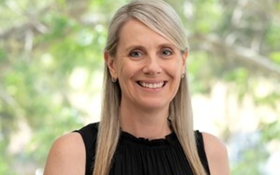 Person with shoulder-length blonde hair, black sleeveless top, yellow flower pin, smiling outdoors.