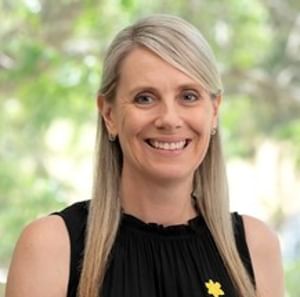Person with shoulder-length blonde hair, black sleeveless top, yellow flower pin, smiling outdoors.