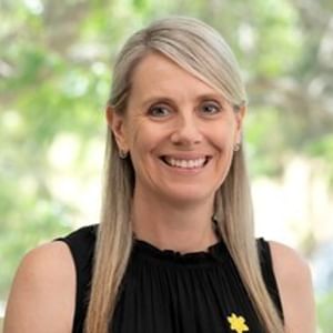 Person with shoulder-length blonde hair, black sleeveless top, yellow flower pin, smiling outdoors.