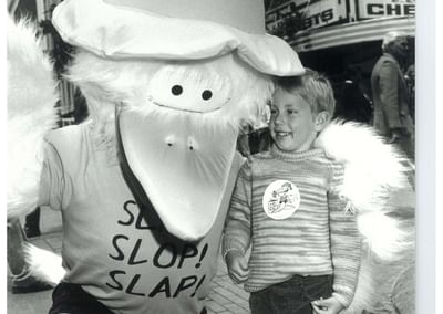 Black-and-white street scene with a large-beaked mascot wearing a hat posing with a smiling child.