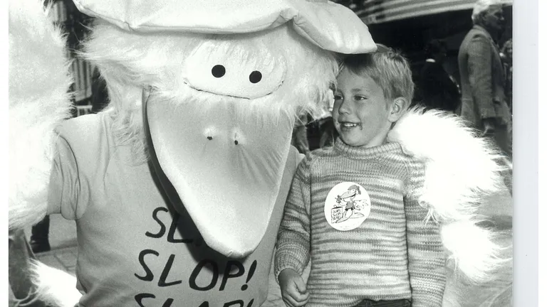 Black-and-white street scene with a large-beaked mascot wearing a hat posing with a smiling child.
