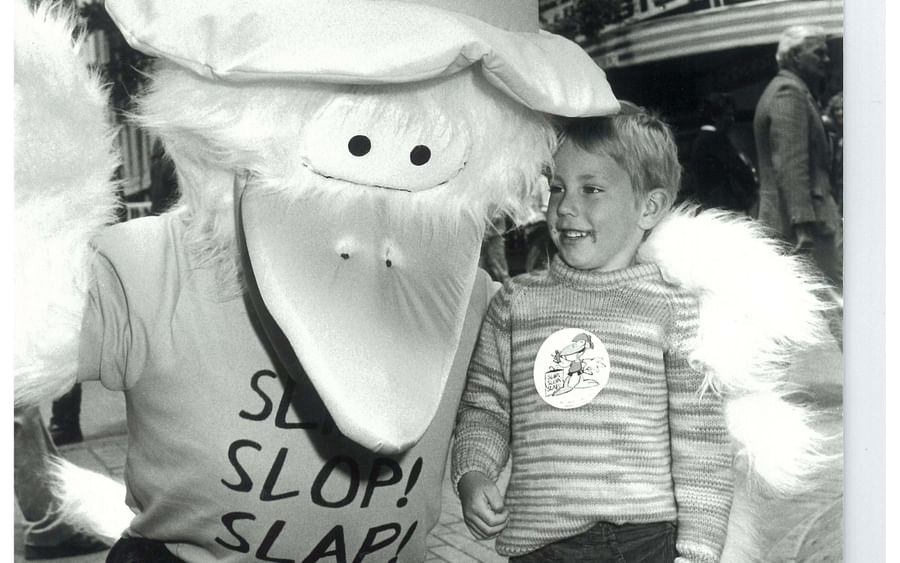Black-and-white street scene with a large-beaked mascot wearing a hat posing with a smiling child.