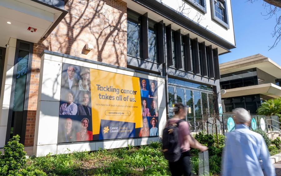 Modern building facade with a large Cancer Council banner reading 'Tackling cancer takes all of us' as pedestrians walk by.