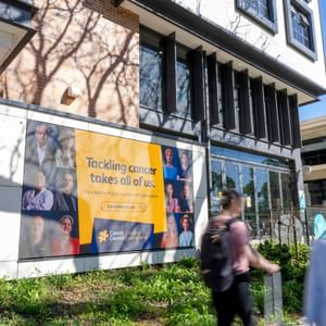 Modern building facade with a large Cancer Council banner reading 'Tackling cancer takes all of us' as pedestrians walk by.