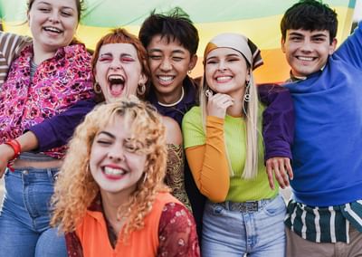 Six diverse people smiling with arms around each other, rainbow flag in the background.