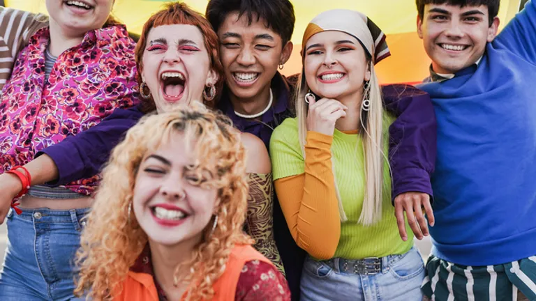 Six diverse people smiling with arms around each other, rainbow flag in the background.