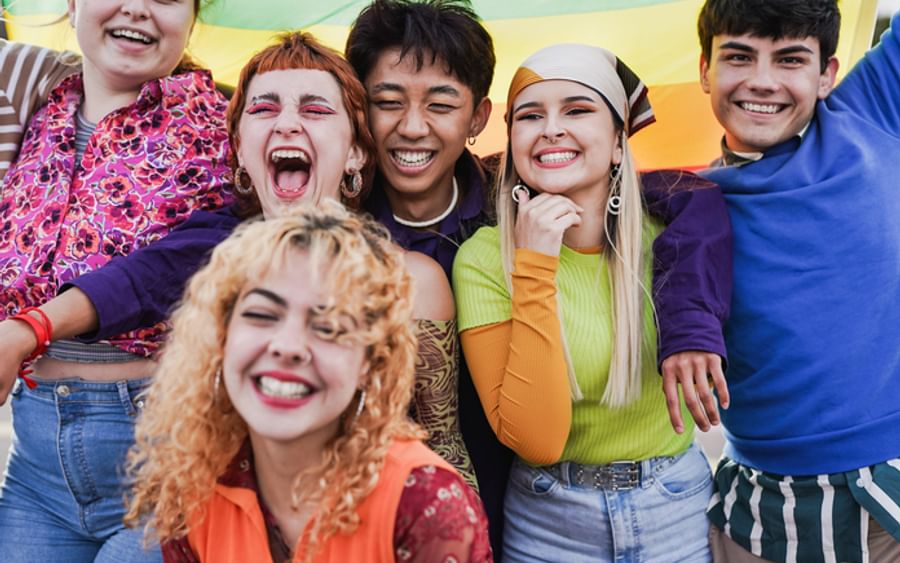 Six diverse people smiling with arms around each other, rainbow flag in the background.