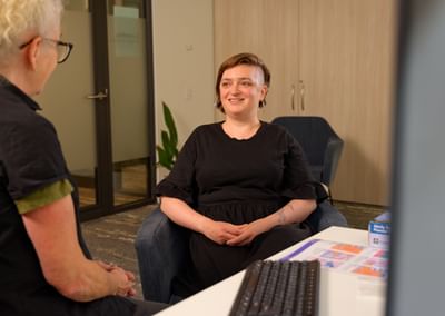 Two people chat in a modern office; one sits smiling in a black dress near a desk.