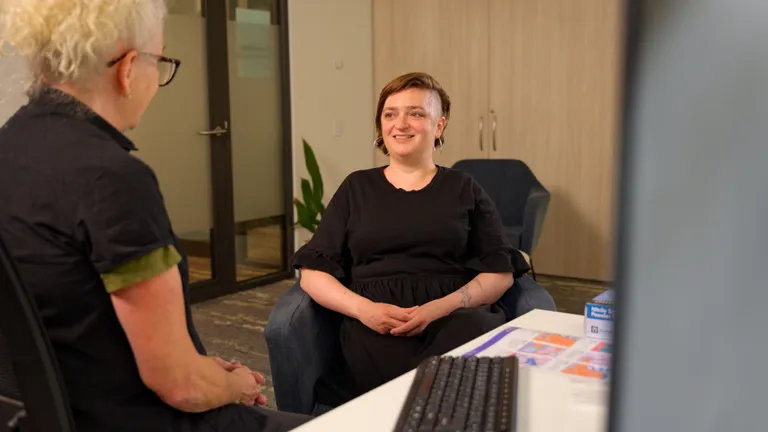 Two people chat in a modern office; one sits smiling in a black dress near a desk.