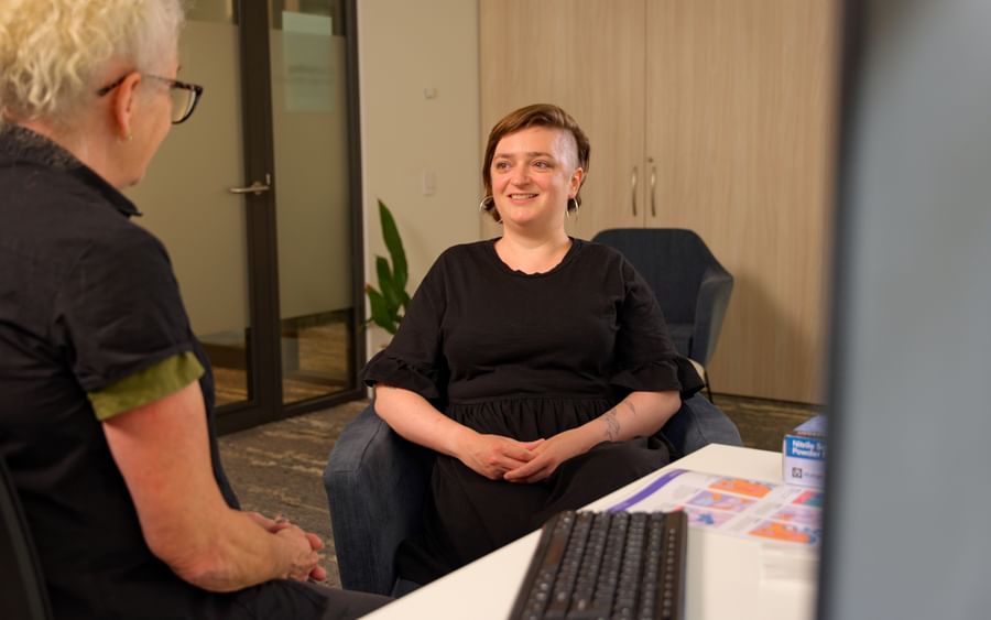 Two people chat in a modern office; one sits smiling in a black dress near a desk.