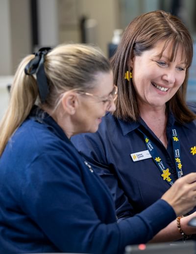 Two staff in navy Cancer Council SA shirts smile at clipboard near a computer, with yellow flowers.