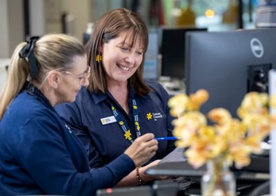 Two staff in navy Cancer Council SA shirts smile at clipboard near a computer, with yellow flowers.