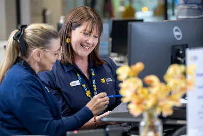 Two staff in navy Cancer Council SA shirts smile at clipboard near a computer, with yellow flowers.