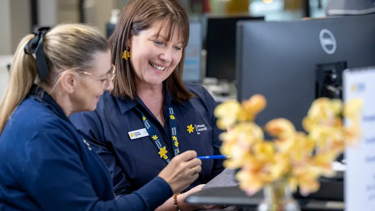 Two staff in navy Cancer Council SA shirts smile at clipboard near a computer, with yellow flowers.