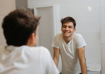 Smiling person in a white T-shirt leans on a bathroom counter, seen in the mirror.