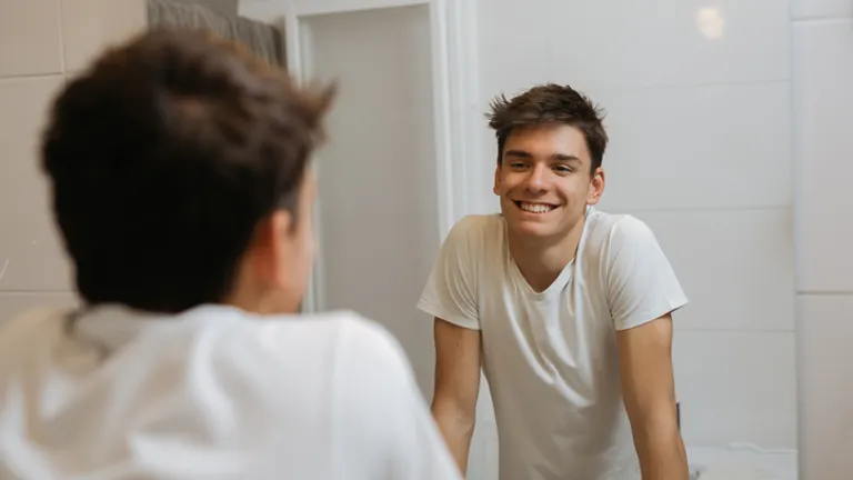 Smiling person in a white T-shirt leans on a bathroom counter, seen in the mirror.
