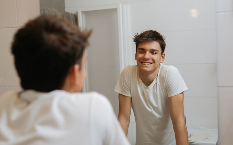 Smiling person in a white T-shirt leans on a bathroom counter, seen in the mirror.