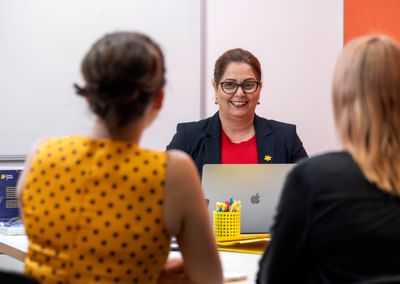 Smiling person in a navy blazer and red top leads a meeting with two colleagues.