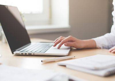 Person typing on a silver laptop at a wooden desk, with a notebook and pen nearby.