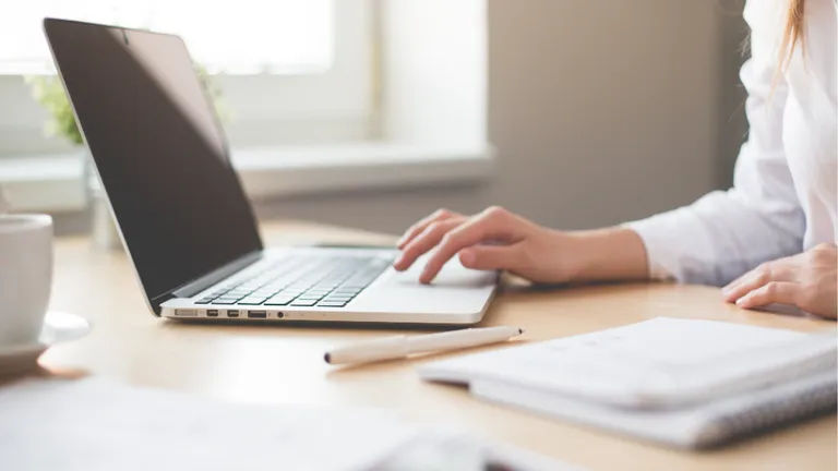 Person typing on a silver laptop at a wooden desk, with a notebook and pen nearby.