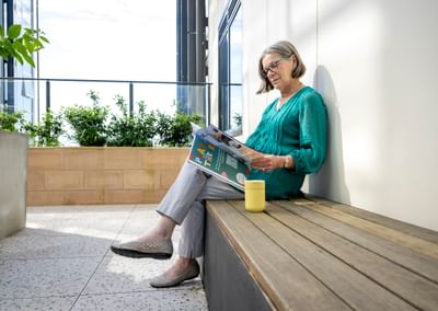 Person sitting on a wooden bench, reading a magazine, wearing a teal top, yellow mug beside.