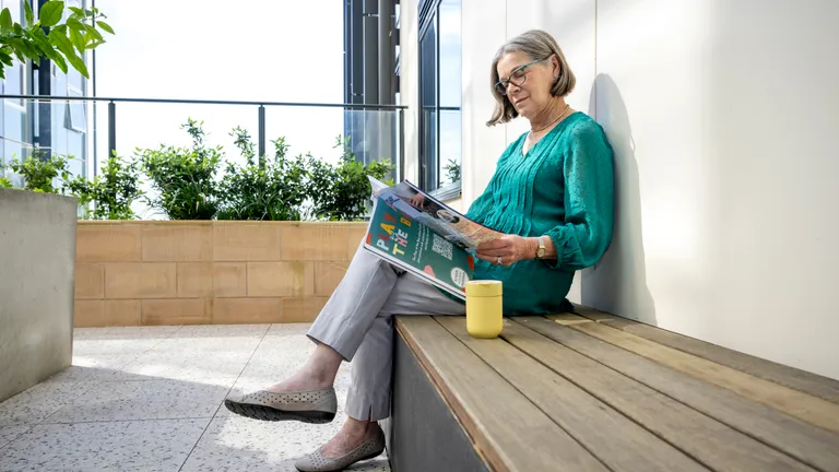 Person sitting on a wooden bench, reading a magazine, wearing a teal top, yellow mug beside.