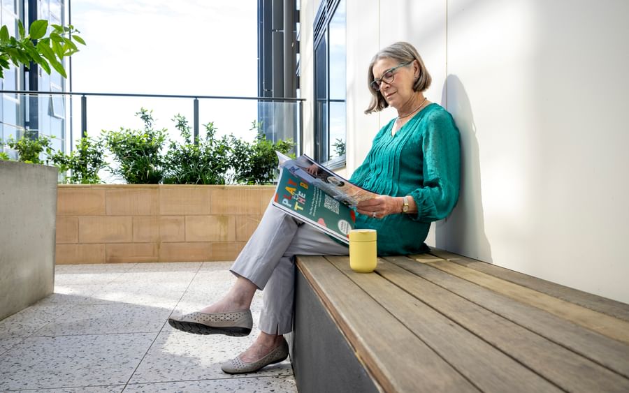 Person sitting on a wooden bench, reading a magazine, wearing a teal top, yellow mug beside.