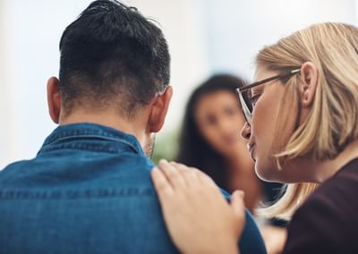 Two people, denim jacket back to camera, blonde glasses wearer touches the other's shoulder.