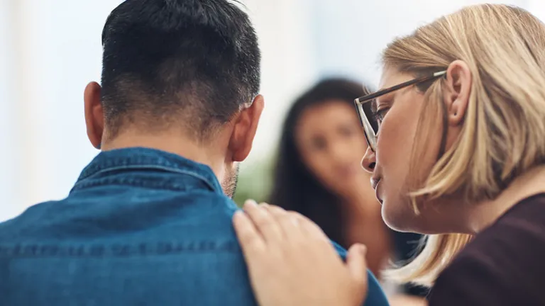 Two people, denim jacket back to camera, blonde glasses wearer touches the other's shoulder.