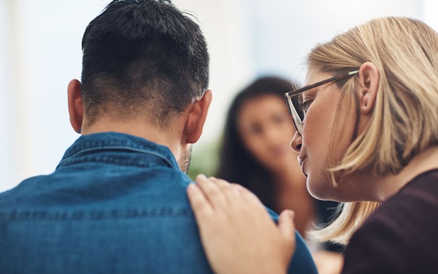 Two people, denim jacket back to camera, blonde glasses wearer touches the other's shoulder.