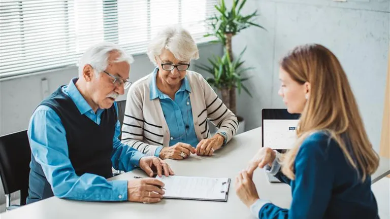 Three people sit around a white table in a bright office, reviewing papers with a laptop.