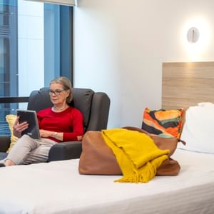 Person sits in a dark armchair by a window, reading a tablet; bed with yellow scarf and bag nearby.