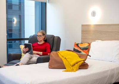 Person sits in a dark armchair by a window, reading a tablet; bed with yellow scarf and bag nearby.