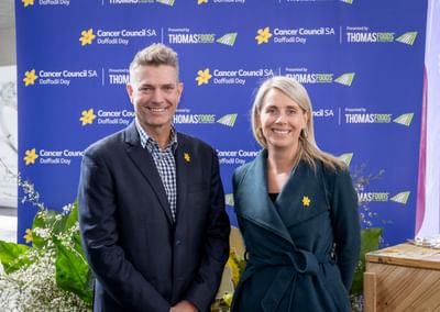 Two people pose in front of a Cancer Council SA Daffodil Day backdrop with yellow daffodil pins.