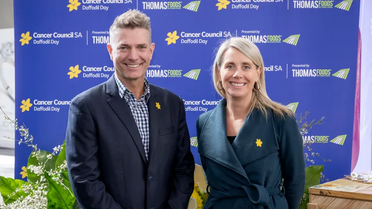 Two people pose in front of a Cancer Council SA Daffodil Day backdrop with yellow daffodil pins.