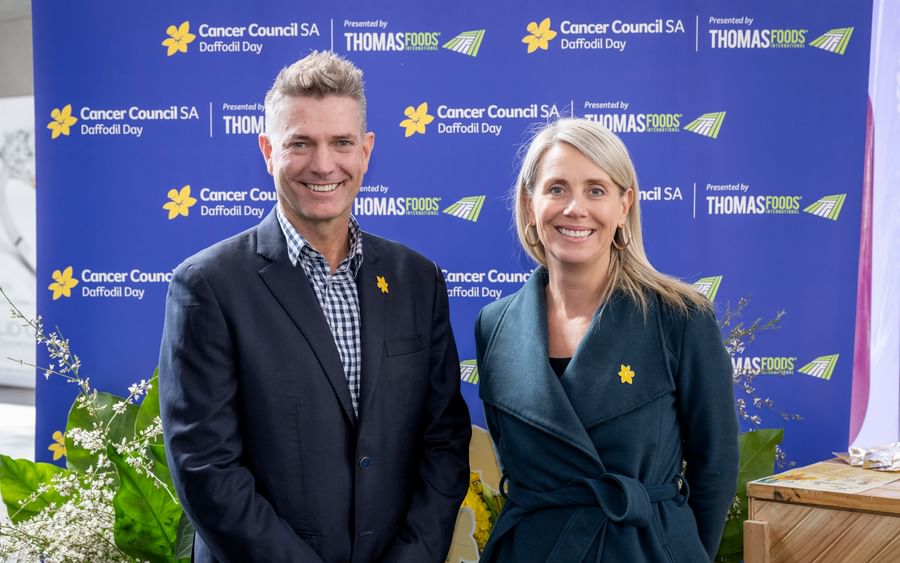 Two people pose in front of a Cancer Council SA Daffodil Day backdrop with yellow daffodil pins.