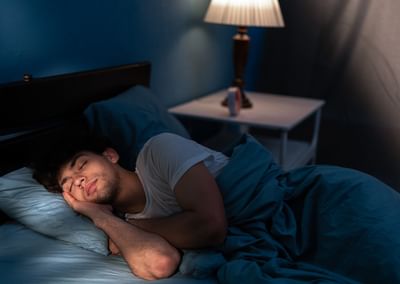 Person sleeping in a blue-lit bedroom on a bed with blue bedding and a lamp on the nightstand.