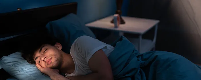 Person sleeping in a blue-lit bedroom on a bed with blue bedding and a lamp on the nightstand.