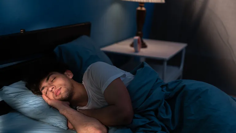 Person sleeping in a blue-lit bedroom on a bed with blue bedding and a lamp on the nightstand.