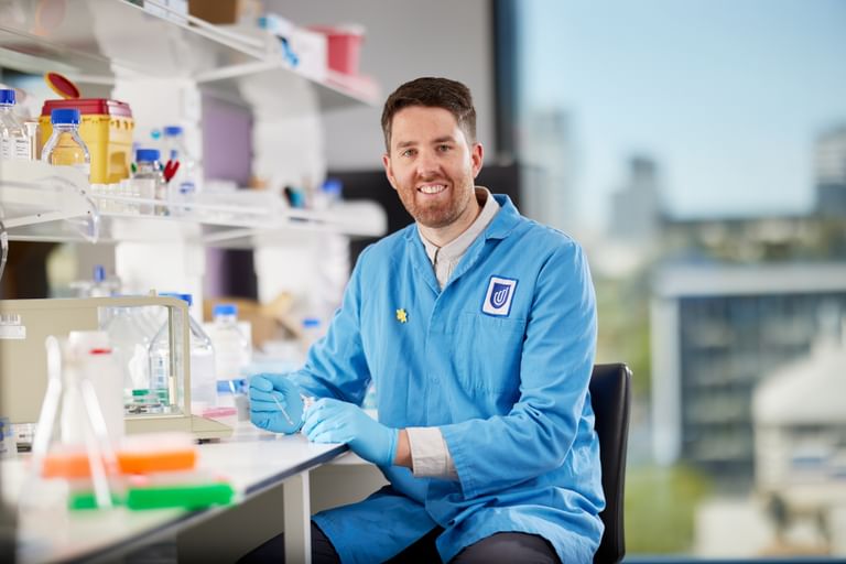 A lab technician in a blue coat and gloves sits at a bench with lab supplies.