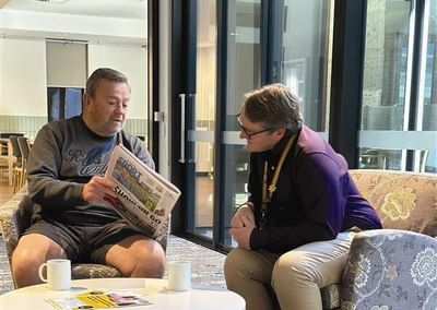 Two people sit in a lounge beside glass doors; one reads a newspaper while the other leans in.