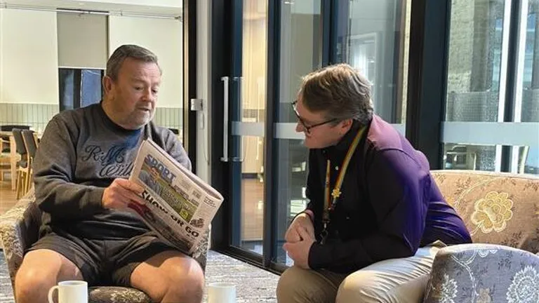 Two people sit in a lounge beside glass doors; one reads a newspaper while the other leans in.