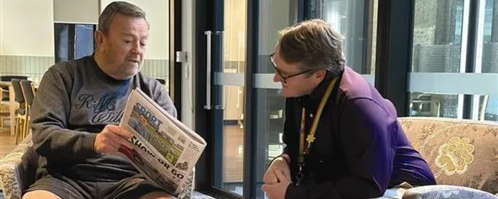 Two people sit in a lounge beside glass doors; one reads a newspaper while the other leans in.